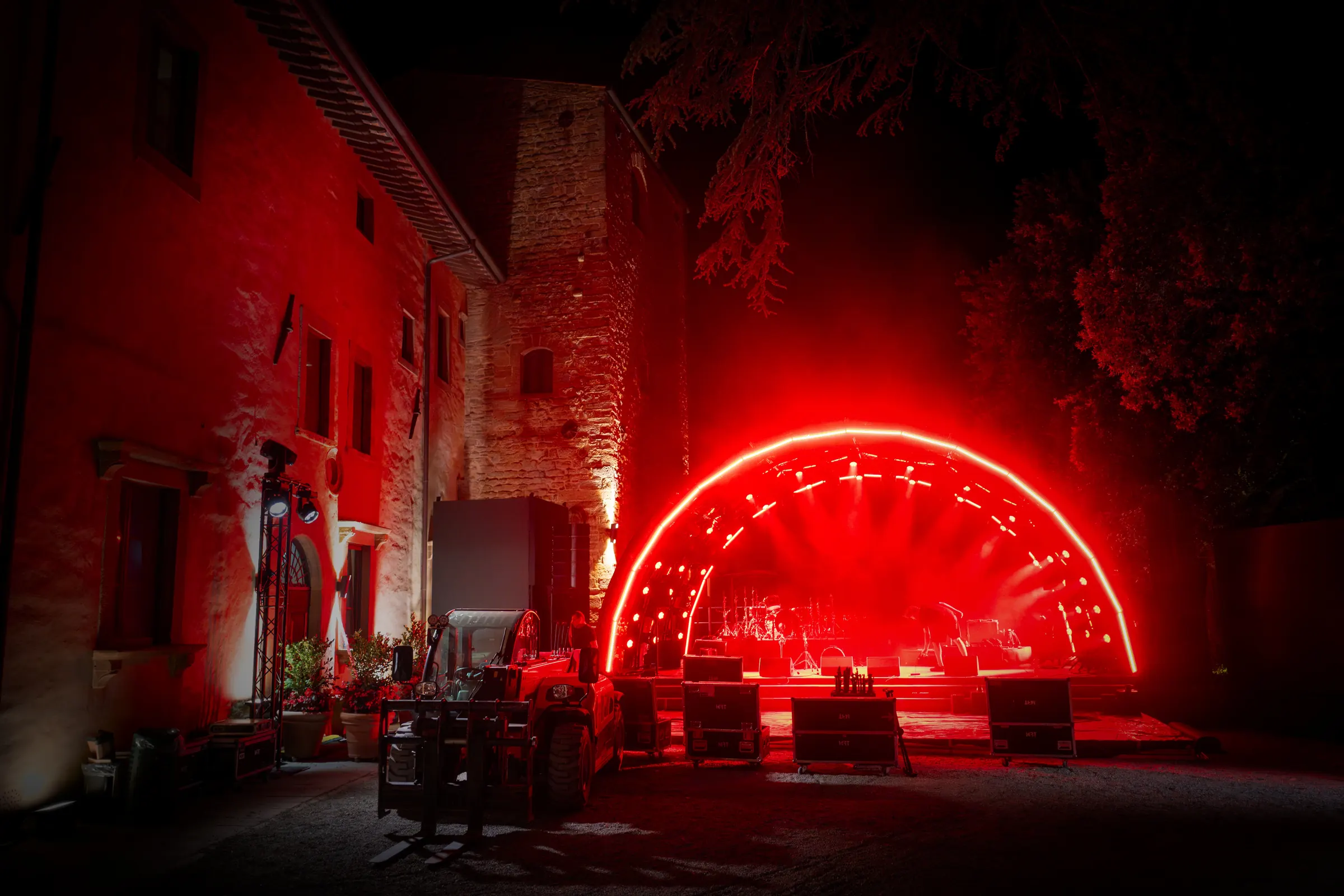 Night setup in a Tuscan courtyard — lit arch stage, road cases and a telehandler before the event